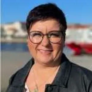 Kvinne med mørkt, kort hår og briller smiler utendørs ved sjø og båter